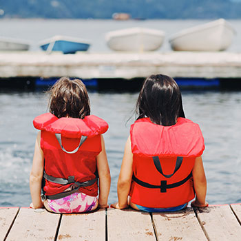 Two young girls with life jackets on sitting on a dock by water